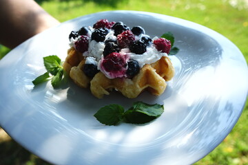 A freshly baked waffle topped with whipped cream and a mix of blackberries and blueberries, presented on a white plate garnished with green mint leaves. 