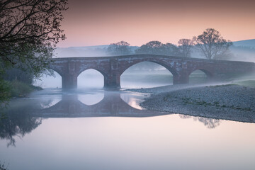 A misty and colourful sunrise during a calm morning looking at Lazonby Eden bridge over the river Eden