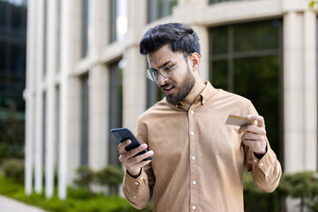 Young man feeling stressed while holding smartphone and credit card in urban setting. Financial concern and frustration evident as he checks digital transaction.
