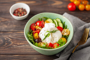 Salad with traditional italian burrata cheese. Burrata ball with cherry tomatoes, cucumber slices, lettuce  and peppercorn in bowl on the table