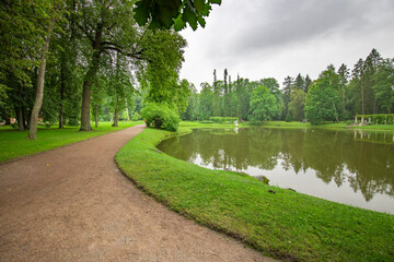 A peaceful pathway winds through greenery, leading to a serene lake. Trees frame the scene under a cloudy sky, inviting wanderers to explore nature's beauty.