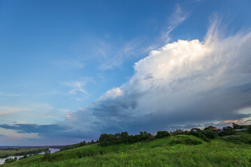 Obraz premium A vast, white cumulus cloud casts a shadow over a green river valley, with a line of trees on the horizon.