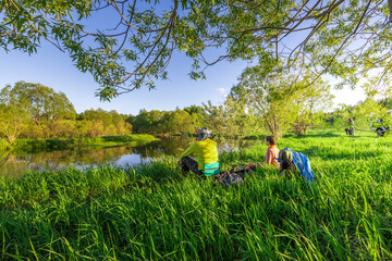 Two enthusiasts relax by a peaceful river, surrounded by vibrant greenery, as sunlight filters through the trees during a calm afternoon in early spring.