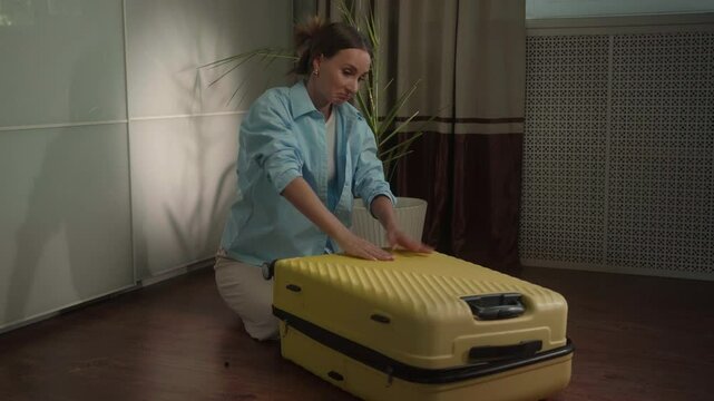 Woman struggles to close her overpacked yellow suitcase while preparing for a trip. Pushing down hard, she battles the challenge of fitting everything inside