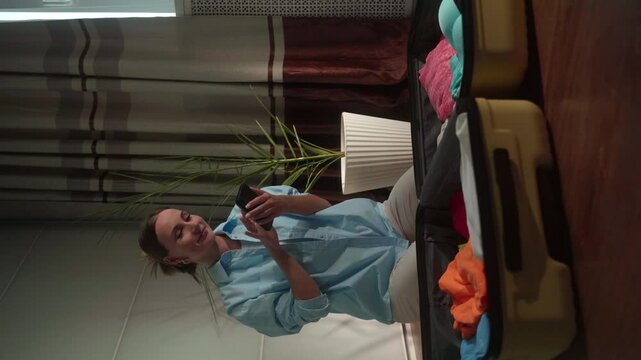 Vertical video: Woman in casual clothing packs a suitcase in a bedroom while happily talking on her phone. Excitement fills the air as she prepares for her upcoming trip
