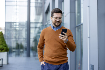 Professional man smiling while reading smartphone outdoors near modern office building during sunny day