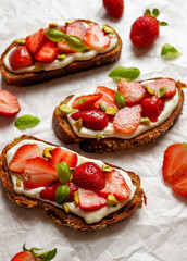 Three slices of open faced sandwich topped with strawberry, greek yogurt, sweet basil leaves, and crushed pistachios, over white background. Close up. Selective focus
