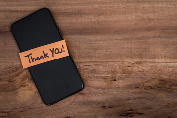 A cell phone with a note on it that says "Thank You". The phone is placed on a wooden surface, and the note is attached to the back of the phone. Concept of gratitude and appreciation