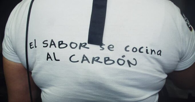 Beautiful on-camera shot of the back of a young Latina chef cooking a grill in a restaurant with a phrase on the T-shirt that reads &ldquo;el sabor se cocina al carb&oacute;n&rdquo;