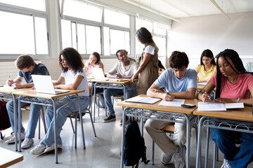In class. Female teacher helping teen male high school student with lesson. Classmates focused on the task at hand.