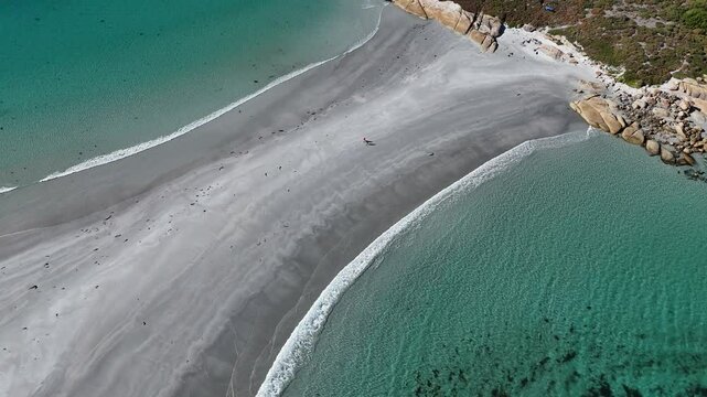 Aerial top down of couple on sandy track between ocean water along shoreline in Tasmania. Visiting Diamond Island during sunny day in summer. Rising drone view.