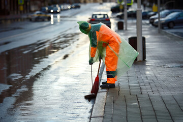 Municipal worker in raincoat cleanup street with broom and dustpan, street workers keep city clean...