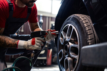 Car mechanist using pneumatic impact wrench to remove the wheel and replace damaged tire in workshop.