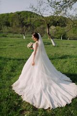 A woman in a white dress is walking through a field. She is holding a bouquet of flowers and a veil. The scene is peaceful and serene, with the woman looking towards the camera