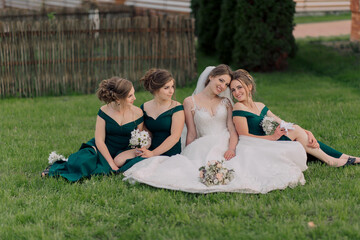 A bride and her bridesmaids are sitting on the grass, posing for a picture. The bride is wearing a...