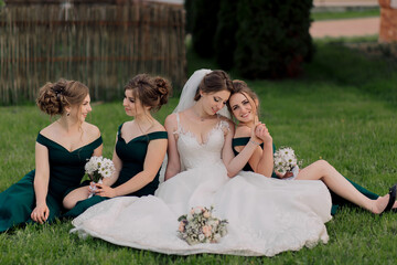 A bride and her bridesmaids are sitting on the grass, with the bride wearing a white dress