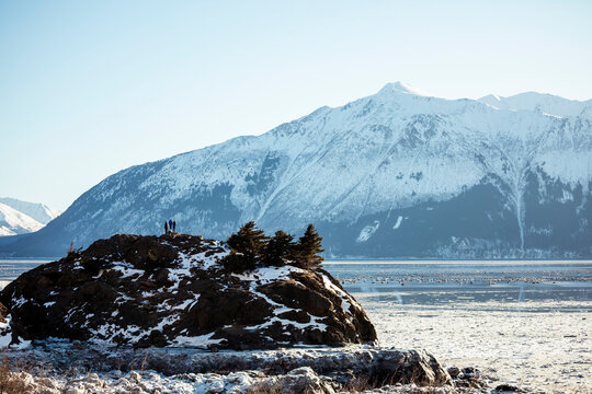 View from behind of three people standing on a rock island looking at the mountains at Beluga Point near Anchorage; Anchorage, Alaska, United States of America