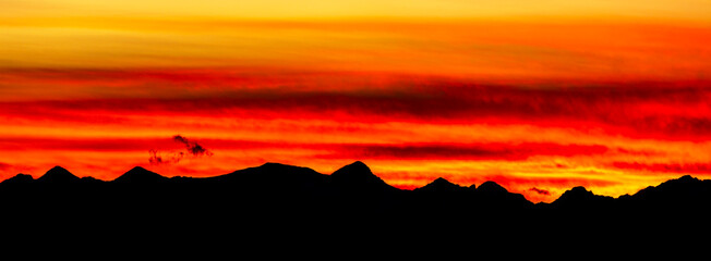 Panoramic of dramatic, colorful red and gold sky with a silhouetted mountain range; Calgary, Alberta, Canada