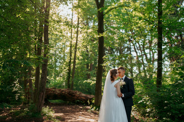 A bride and groom are standing in a forest, surrounded by trees. The bride is wearing a white dress and the groom is wearing a suit. They are both smiling and looking at the camera