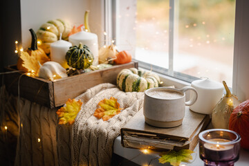 Mug of coffee standing next to the window with candles, pumpkins, book and warm blanket. Cozy home atmosphere in sunny autumn day. Cozy fall composition.