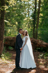 A bride and groom are standing in a forest, with the bride wearing a white dress and the groom wearing a suit. They are holding hands and kissing, surrounded by trees and sunlight