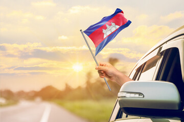 Waving the Cambodia flag against the sunrise or sunset from a car driving along a country road. Holding the Cambodia flag, traveling by car, on a weekend trip.