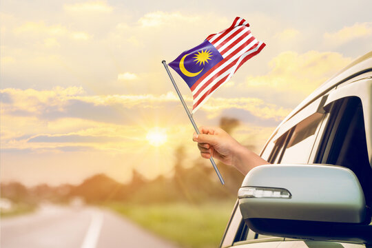 Waving the Malaysia flag against the sunrise or sunset from a car driving along a country road. Holding the Malaysia flag, traveling by car, on a weekend trip.