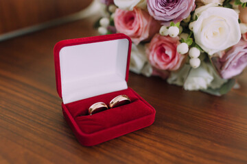 A red box with two gold wedding rings inside. The box is placed on a wooden table next to a bouquet of pink flowers