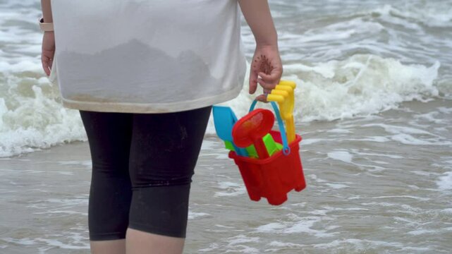 Indian woman in white shirt standing on sea shore, beach looking at waves, holding a colorful sand castle making kit toy at baga beach in goa India