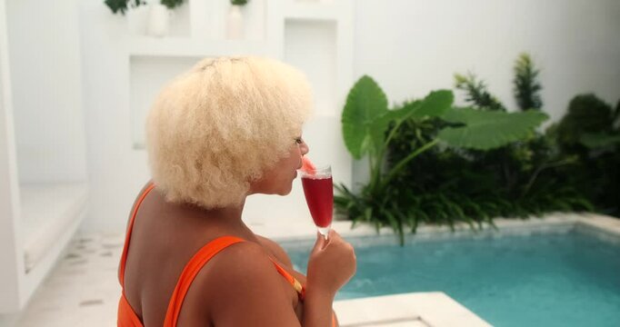 A Young African American curvy woman in an orange bikini drinking watermelon cocktail from a glass sitting by the swimming pool. Summer vacation in luxury hotel in tropics. Summer drinks.