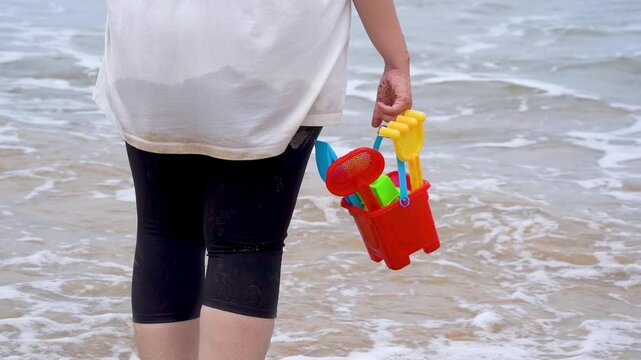 Slow motion shot Indian woman in white shirt standing on sea shore, beach looking at waves, holding a colorful sand castle making kit toy at baga beach in goa India
