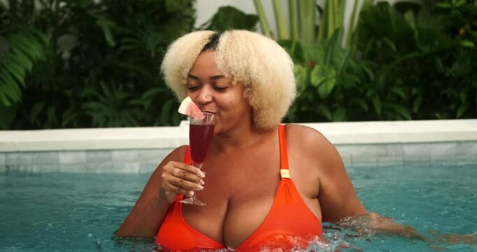 Young African American woman with big breasts in orange bikini enjoying relaxation in blue water pool. She holds glass with red cocktail and watermelon slice. Cooling off in heat. Tropical vacation.