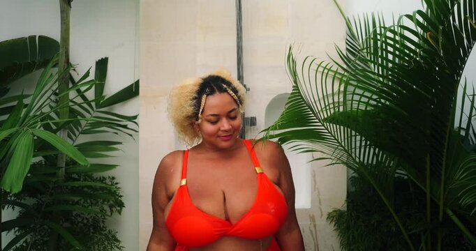 Portrait of young African American curvy woman with big breasts in bikini standing underwater in outdoor tropical shower. Palm trees and other green plants grow in bathroom. Self care and cleansing.