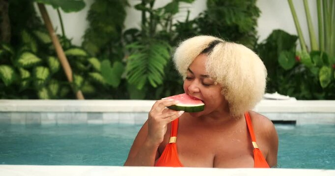 Young African American sexy plus size woman with big boobs in bikini enjoying relaxing in the pool and eating a piece of juicy watermelon. Fresh summer fruits during vacation. Detox diet.