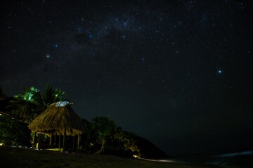 Stars and the Milky Way above a small hut on the beach. Fiji nighttime beach scene. Stunning night sky over the Yasawa Islands, Fiji. Palm trees, stars and the beach in the night. 