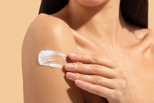 Woman spreading cream on her shoulder, taking care of herself, swatch of cream, beige background, closeup shot