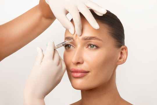 Beautician making hyaluronic acid injection for glabella facial rejuvenation procedure, woman getting cosmetic injection between eyebrows over white studio background