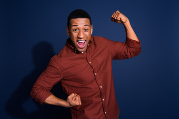 Photo of lucky cool guy dressed brown shirt rising fists emtpy space isolated blue color background