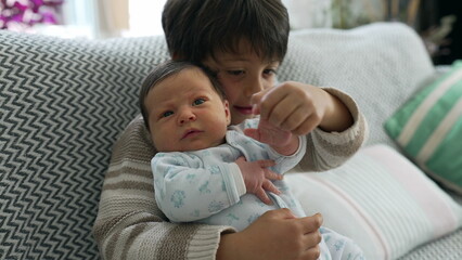 Young boy holding newborn baby, both sitting on a sofa. baby looks content while the boy shows curiosity, capturing a precious family moment of sibling bonding