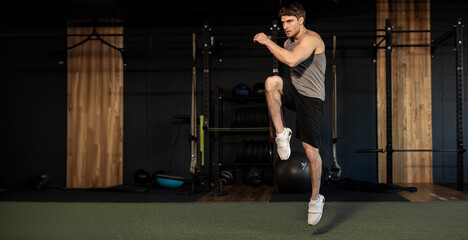 Energetic man doing fitness exercise, warming up and lifting leg, full length shot, gym interior