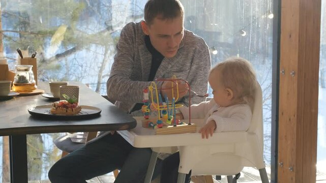 Father lovingly feeds his baby daughter a waffle dessert while enjoying a warm, inviting atmosphere in a cafe surrounded by a winter forest
