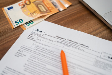 Employee filling out a W-4 tax form with euro banknotes and laptop on wooden desk. Financial documentation and tax preparation concept 