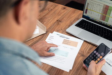 Man reviewing utility bills at home while using a smartphone calculator. Personal finance...