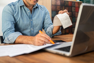 Middle-aged man calculating taxes at a wooden desk with a laptop and holding receipts. Personal...