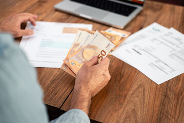 Man handling euro bills while reviewing utility expenses and household finances. Emphasis on financial planning, tax calculations, and managing payments online 