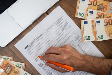 Fototapeta premium Hand of a man filling out a 1040 tax form with euros and a laptop on a wooden desk. Tax preparation and financial documentation concept, suitable for accounting and international finance themes 