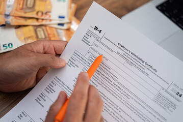 Freelancer meticulously reads W-4 form for employer to know exactly sum of money federal tax to withhold from earnings at dining table with laptop and euro bills