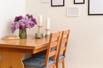 Vase with beautiful lilac flowers on table, chairs and blank pictures in interior of light living room