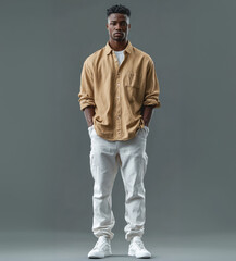 Obraz premium A young African American man in a casual outfit with a tan button-up shirt and white pants stands confidently with hands in pockets against a plain gray background, exuding a relaxed and modern vibe.