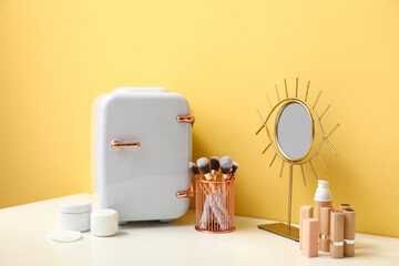 Portable fridge with different makeup products and mirror on dressing table near beige wall in room